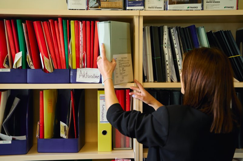 Pracownica biura New-House sięgająca po segregator z dokumentacją inwestycyjną. Zdjęcie ilustruje proces kompletowania dokumentów do pozwolenia na budowę i warunków zabudowy, który generalny wykonawca realizuje w imieniu klienta, zdejmując z niego ciężar biurokracji.